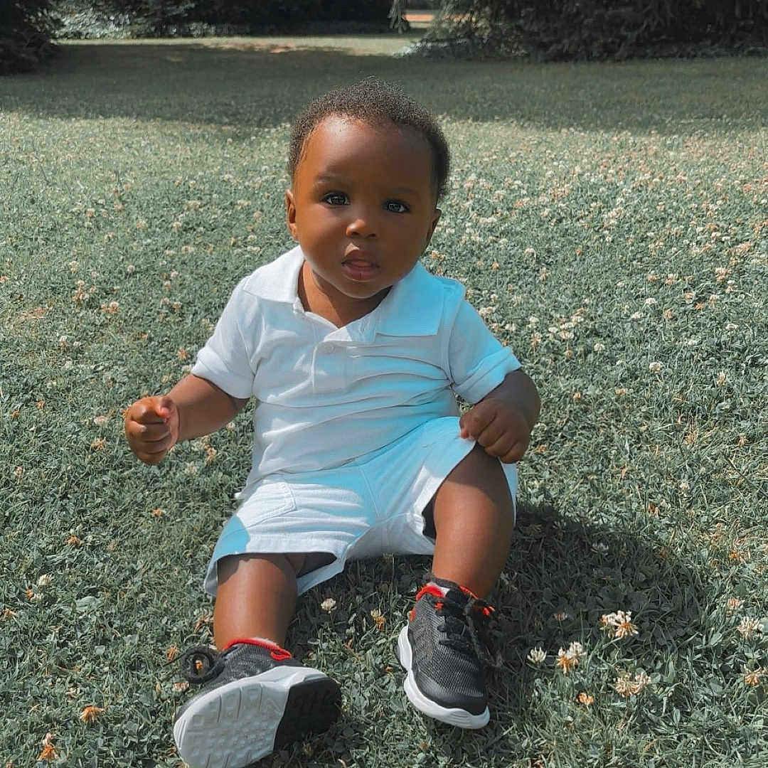 Hiram Treh a rejoint le concours — aidez-le/la à gagner de superbes lots ! black_shoes, casual_clothing, child, curious, cute, daylight, flowers, grass, nature, outdoor, park, portrait, red_accents, sitting, sneakers, summer, toddler, trees, white_clothing, young_child