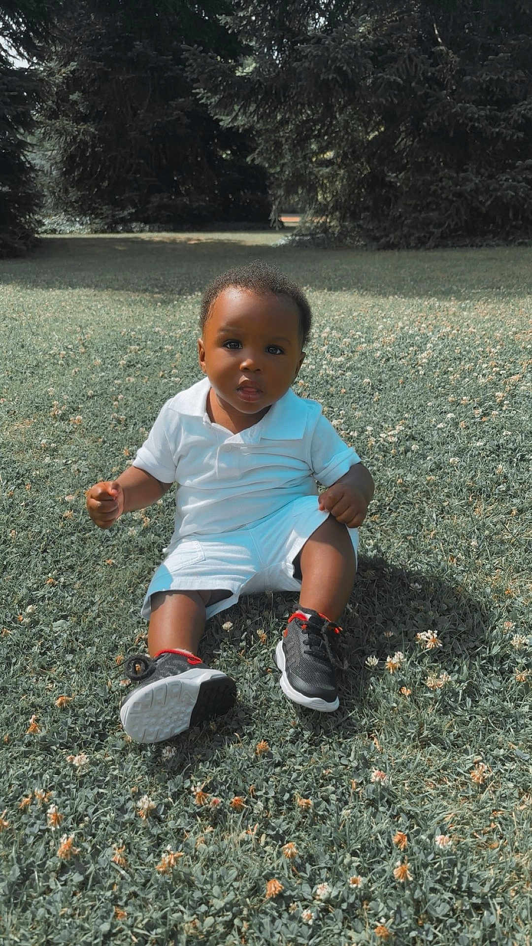 Hiram Treh a rejoint le concours — aidez-le/la à gagner de superbes lots ! toddler, child, grass, outdoor, nature, trees, flowers, white_clothing, sneakers, black_shoes, red_accents, sitting, curious, portrait, daylight, young_child, casual_clothing, park, summer, cute