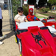 Nirvair is registered to the contest to win money with this photo: child, kid, spiderman, ride, kiddie_ride, toy_car, red_car, outdoor, sunny_day, park, fence, people, adult, shoes, concrete, tree, greenery, flowers, sidewalk, costume