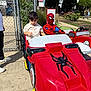 child, kid, spiderman, ride, kiddie_ride, toy_car, red_car, outdoor, sunny_day, park, fence, people, adult, shoes, concrete, tree, greenery, flowers, sidewalk, costume