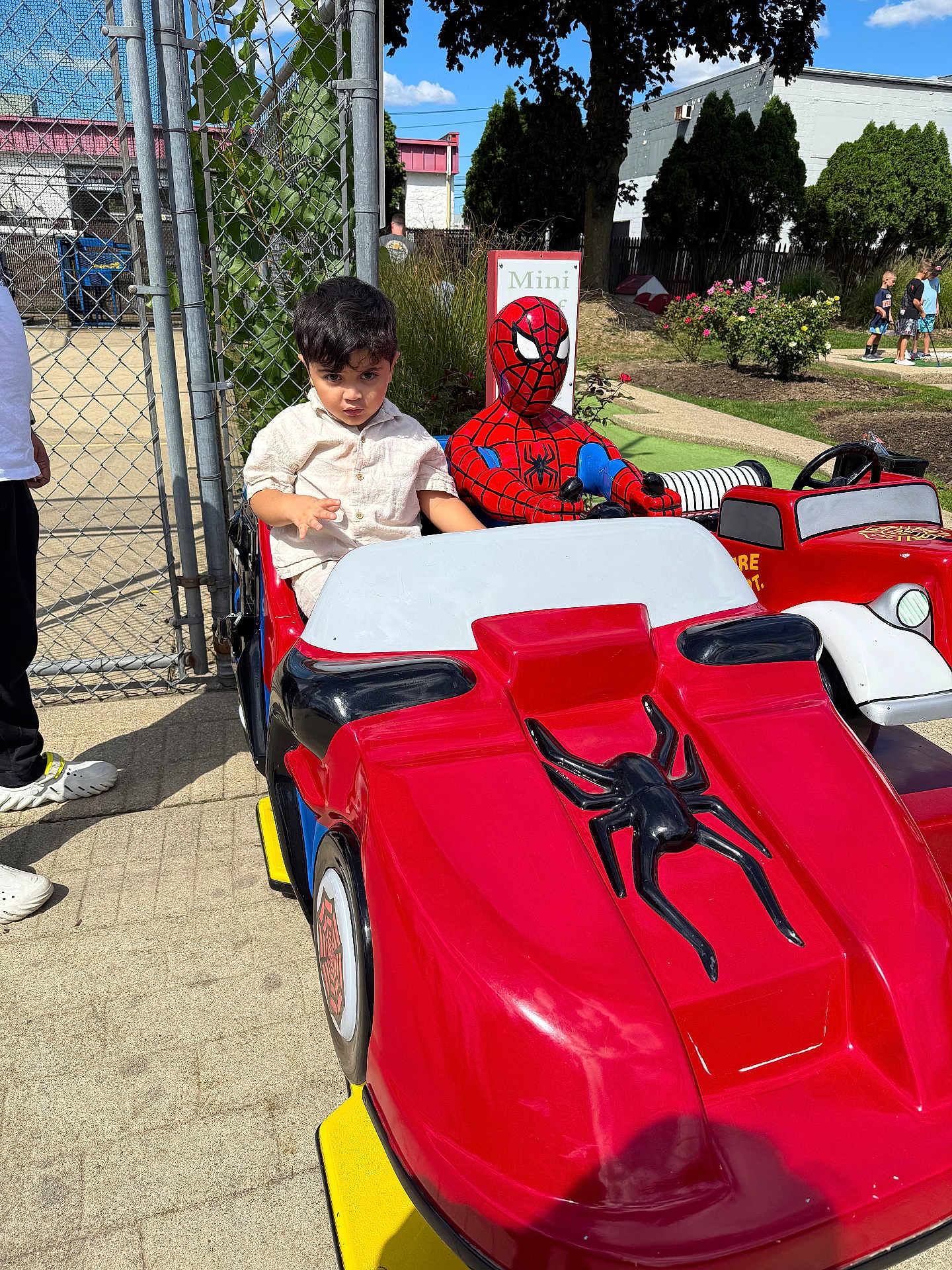 Nirvair is registered to the contest to win money with this photo: child, kid, spiderman, ride, kiddie_ride, toy_car, red_car, outdoor, sunny_day, park, fence, people, adult, shoes, concrete, tree, greenery, flowers, sidewalk, costume