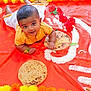 baby, child, yellow_shirt, red_cloth, rice_pattern, roses, flatbread, marigold_flowers, decorative, curious, smiling, lying_down, indoors, celebration, festive, portrait, cute, person, infant, colorful