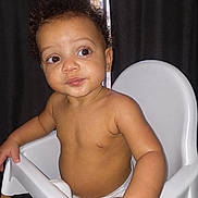 Chandler is registered to the contest to win money with this photo: baby, child, infant, high_chair, portrait, sitting, indoors, curly_hair, brown_skin, bare_chested, diaper, face, eyes, looking_at_camera, serious_expression, white_chair, curtains, window, flash_photography, home