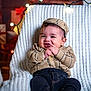 baby, child, smile, plaid_shirt, cap, socks, dark_pants, knitted_blanket, chair, fairy_lights, cozy, indoor, portrait, cute, happy, infant, festive, warm_lighting, hands, playful