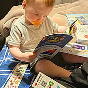 Kayson is registered to the contest to win money with this photo: toddler, child, pacifier, book, reading, blanket, indoors, education, learning, sitting, clothing, toy, floor, person, light, hair, face, curtain, background, colorful