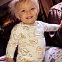 toddler, child, smiling, blonde_hair, indoor, pajamas, cushion, patterned, wooden_furniture, cozy, warm_light, face, person, happy, cute, home, plush, sitting, blanket, young_child
