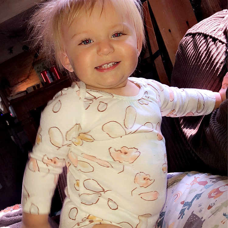 Maevyn is registered to the contest to win money with this photo: blanket, blonde_hair, child, cozy, cushion, cute, face, happy, home, indoor, pajamas, patterned, person, plush, sitting, smiling, toddler, warm_light, wooden_furniture, young_child