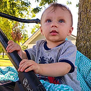 Axel is registered to the contest to win money with this photo: baby, child, stroller, outdoor, tree, sunlight, blanket, grass, park, curious, hands, face, blue_eyes, shirt, casual_clothing, nature, daytime, expression, person, infant