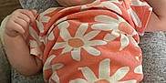Jupiter is registered to the contest to win money with this photo: baby, infant, onesie, orange_clothing, daisy_pattern, soft_surface, grey_fabric, wooden_floor, curious_expression, small_hands, lying_down, indoor, person, child, cute, portrait, casual, comfortable, young, innocent