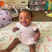 Rumani is registered to the contest to win money with this photo: baby, child, clothing, dress, face, floor, furniture, hair_bows, happy, indoors, laundry_basket, numbers, person, playmat, room, sitting, smiling, storage_drawers, toddler, toys