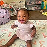 baby, child, clothing, dress, face, floor, furniture, hair_bows, happy, indoors, laundry_basket, numbers, person, playmat, room, sitting, smiling, storage_drawers, toddler, toys