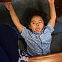 child, curly_hair, blue_shirt, tongue_out, arms_up, indoor, carpet, wooden_table, playful, person, smiling, cheeky, furniture, floor, casual_clothing, happy, portrait, young, fun, relaxed