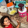 child, smiling, curly_hair, kitchen, countertop, baking, brown_sugar, cocoa, eggs, coffee, mixing_bowl, whisk, spatula, container, floor, cabinet, food, ingredient, happy, person