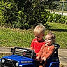 baby, backyard, blue_vehicle, children, driving, family, fence, garden, grass, orange_clothing, outdoor, playtime, red_shirt, seated, siblings, smiling, sunlight, toddlers, toy_car, toy_jeep