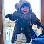 baby, infant, child, smiling, winter_clothing, hat, mittens, snow, window, wooden_frame, jacket, coat, adult_hand, indoor, portrait, happy, playful, cold_weather, balcony_background, building_exterior