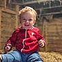 barn, blue_pants, casual_clothing, child, childhood, curly_hair, cute, footwear, happy, hay, indoor, natural_light, playful, portrait, red_jacket, rustic, sitting, smiling, toddler, wooden_walls