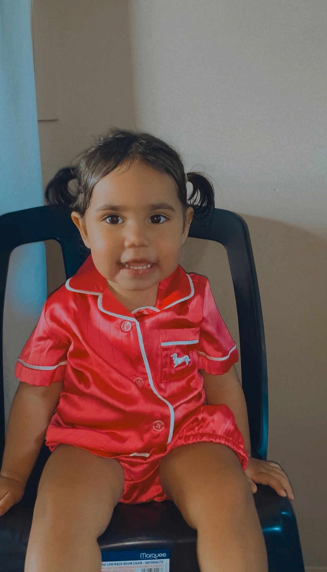 Kytaiviah is registered to the contest to win money with this photo: black_chair, buttoned_shirt, chair, child, clothing, cute, face, happy, indoor, person, pigtails, portrait, red_pajamas, seated, shorts, skin, smile, toddler, wall, young_child