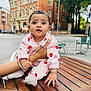 Samriddha joined the competition — help win amazing prizes! baby, child, infant, pink_clothing, strawberry_pattern, table, hand, outdoor, urban, building, brick_building, street, greenery, trees, chair, footwear, person, face, cute, portrait