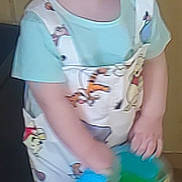 Joey is registered to the contest to win money with this photo: toddler, child, smiling, barefoot, toy, bucket, overalls, cartoon_prints, curly_hair, indoor, floor, happy, clothing, person, portrait, playful, light_blue, standing, young_child, cute