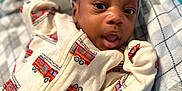 Jelani is registered to the contest to win money with this photo: baby, infant, onesie, fire_truck, hat, blanket, plush_toy, bed, portrait, face, clothing, cute, child, lying_down, comfort, expression, cozy, indoor, soft, closeup