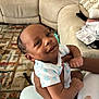 Jelani is registered to the contest to win money with this photo: baby, child, smiling, onesie, blue_stars, hands, adult, living_room, couch, rug, patterned_rug, carpet, person, infant, holding, happy, indoor, face, skin, cozy