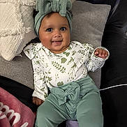 Milah is registered to the contest to win money with this photo: baby, child, smiling, headband, bow, long_sleeve_shirt, patterned_clothing, pants, couch, pillows, blanket, indoor, happy, infant, person, seated, cute, face, cozy, soft