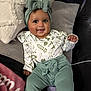 baby, child, smiling, headband, bow, long_sleeve_shirt, patterned_clothing, pants, couch, pillows, blanket, indoor, happy, infant, person, seated, cute, face, cozy, soft