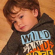 Tatum joined the competition — help win amazing prizes! child, boy, portrait, lying_down, wooden_floor, indoors, brown_hair, brown_eyes, sweatshirt, graphic_shirt, clothing, face, head, eyes, looking_at_camera, calm, thoughtful, closeup, warm_lighting, photography