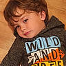 child, boy, portrait, lying_down, wooden_floor, indoors, brown_hair, brown_eyes, sweatshirt, graphic_shirt, clothing, face, head, eyes, looking_at_camera, calm, thoughtful, closeup, warm_lighting, photography