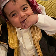 Saeed is registered to the contest to win money with this photo: baby, child, smiling, traditional_clothing, headscarf, golden_embroidery, couch, yellow_cushion, hand, face, happy, indoors, person, seated, cute, portrait, clothing, young_child, fabric, home