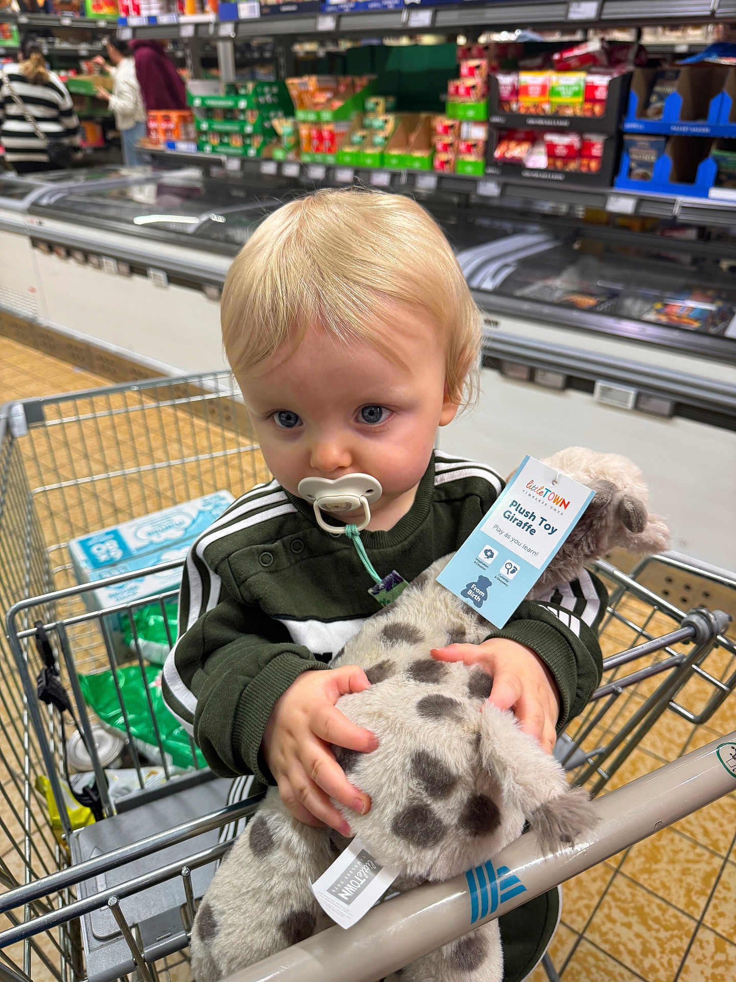 Noah joined the competition — help win amazing prizes! child, toddler, shopping_cart, plush_toy, giraffe, pacifier, blond_hair, blue_eyes, clothing, green_jacket, hand, supermarket, aisle, products, floor, metal, person, toy, holding, shopping