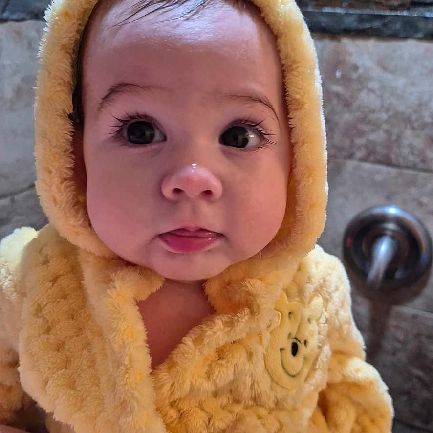 Zulema is registered to the contest to win money with this photo: baby, big_eyes, child, closeup, curious_expression, cute, face, fluffy_clothing, hooded_jacket, indoor, infant, metal_fixture, person, portrait, skin, soft_texture, tiled_wall, tongue_out, warm_clothing, yellow_hood