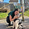 woman, dog, outdoor, sunlight, hug, affection, pet, grass, pavement, playground, fence, tree, house, smile, black_and_white_dog, casual_clothing, happy, leisure, daytime, nature