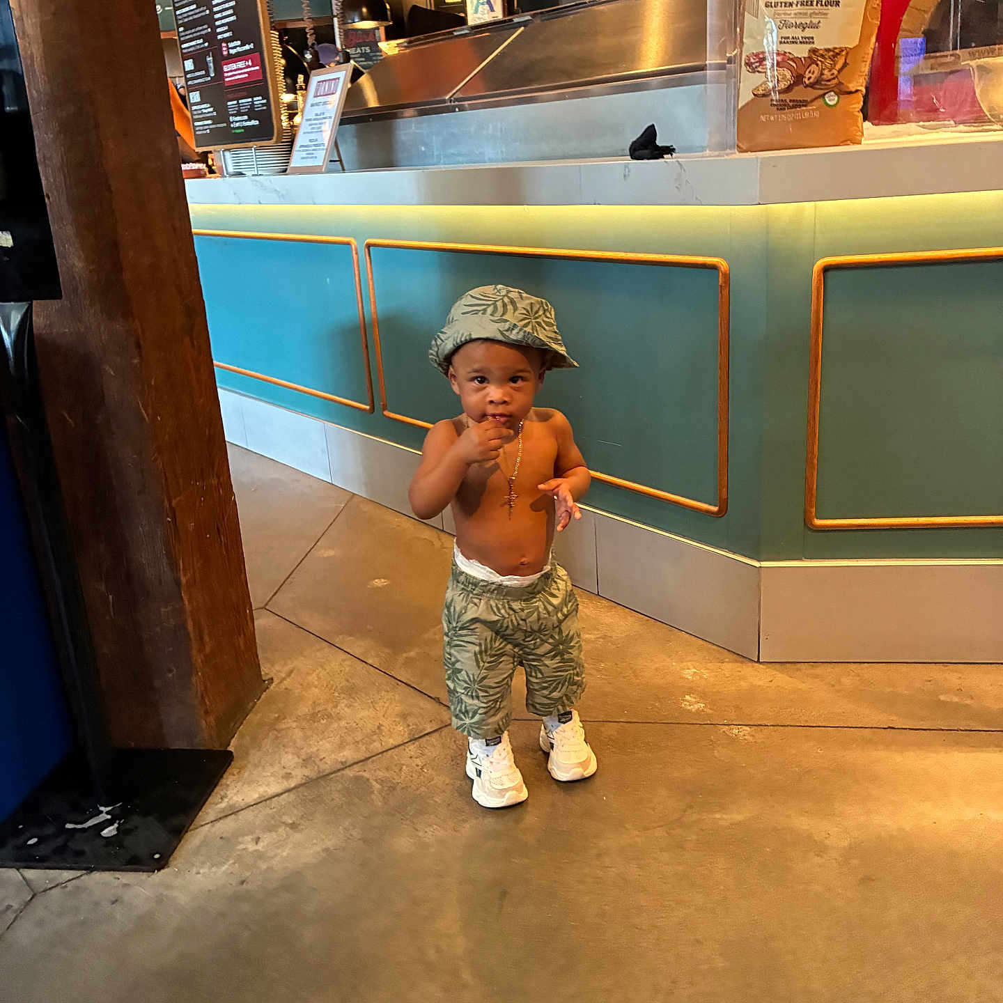 Leslie is registered to the contest to win money with this photo: toddler, child, hat, shorts, sneakers, gold_chain, indoor, floor, counter, cafe, curious, standing, fashion, baby, person, clothing, footwear, concrete_floor, menu, casual