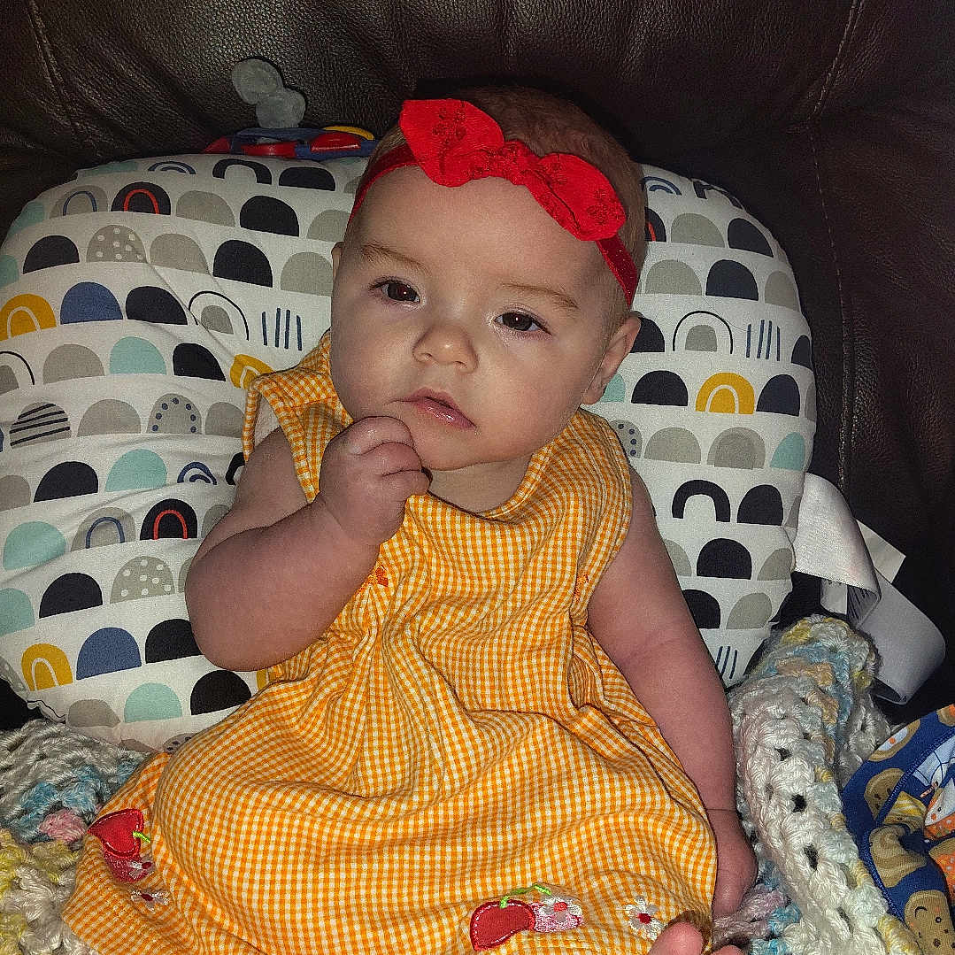 Stirling joined the competition — help win amazing prizes! baby, child, infant, dress, headband, red_bow, yellow_dress, crocheted_blanket, cushion, leather_sofa, foot, hand, face, seated, indoors, portrait, curious, cute, soft_toys, colorful