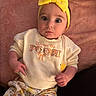 baby, child, infant, portrait, sitting, lying_down, yellow_bow, headband, wide_eyes, serious_expression, pink_blanket, floral_clothing, sweater, baby_outfit, indoor, cozy, close_up, face, text_on_clothing, photography