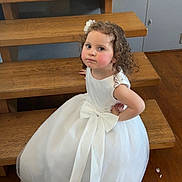 Alba a rejoint le concours — aidez-le/la à gagner de superbes lots ! child, girl, white_dress, curly_hair, bow, stairs, wooden_floor, rosy_cheeks, posing, indoor, person, cute, fashion, portrait, smile, young_child, formal_wear, floor, wall, flower_hair_accessory
