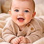 baby, smiling, blue_eyes, knitted_sweater, wooden_buttons, cozy, blanket, soft_texture, cute, happy, infant, portrait, child, laying_down, hands_together, close_up, indoor, warm_colors, skin, face