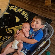 Paxton is registered to the contest to win money with this photo: baby, baby_holding, baby_rocker, blue_shirt, casual_clothing, child, couch, diaper, family, footwear, indoor, living_room, pacifier, person, rug, seat, sibling, smile, table, wooden_floor