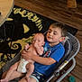 baby, child, sibling, baby_rocker, blue_shirt, diaper, living_room, rug, wooden_floor, couch, table, footwear, baby_holding, family, indoor, smile, person, seat, pacifier, casual_clothing