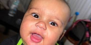 Wysdom is registered to the contest to win money with this photo: baby, child, face, smile, mouth_open, black_shirt, green_trim, red_text, indoor, blurred_background, table, bottle, chair, furniture, person, head, eyes, happy, infant, portrait