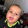 baby, child, face, smile, mouth_open, black_shirt, green_trim, red_text, indoor, blurred_background, table, bottle, chair, furniture, person, head, eyes, happy, infant, portrait