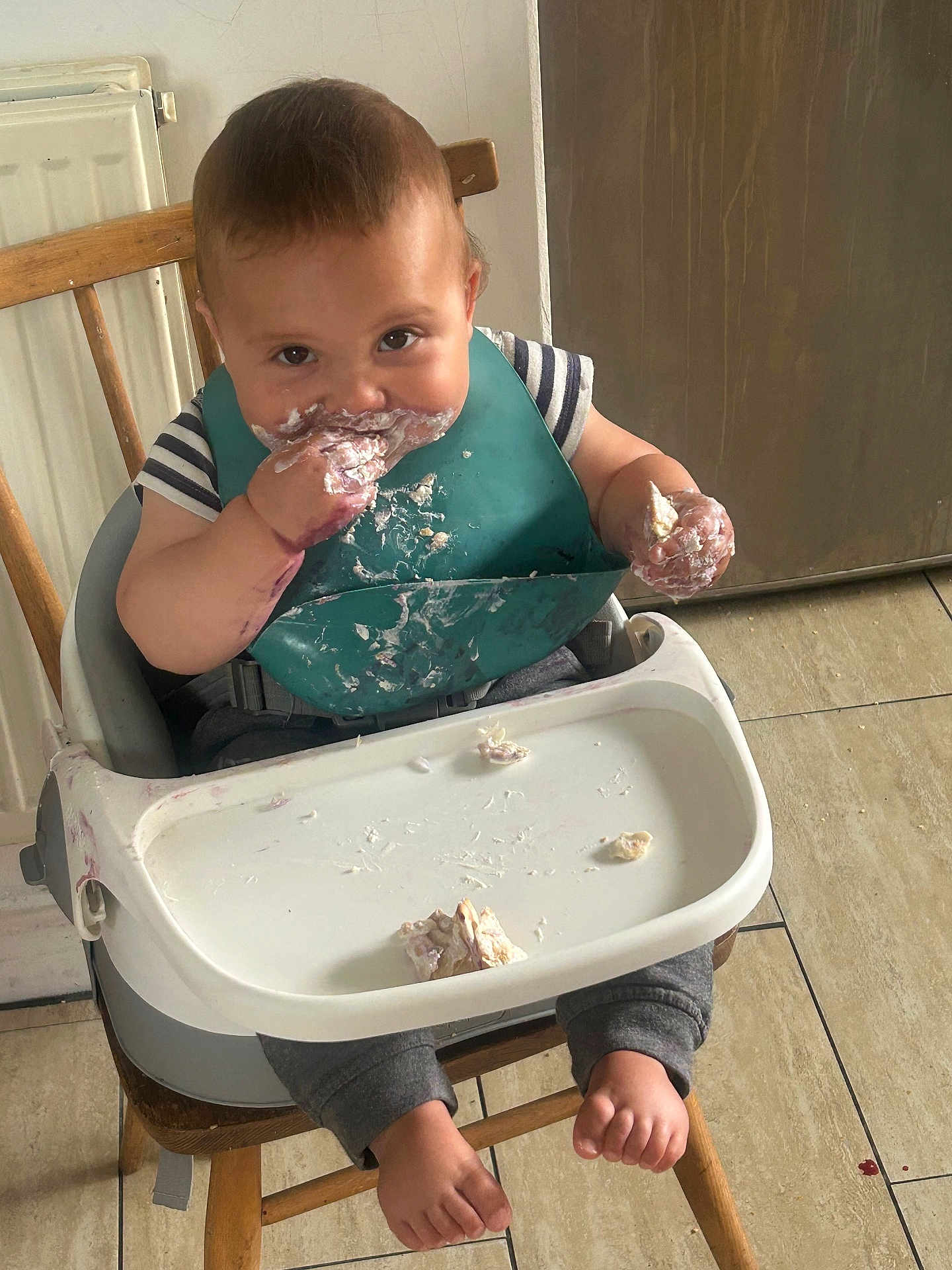 Ezra joined the competition — help win amazing prizes! toddler, child, high_chair, messy, food, bib, eating, smiling, bare_feet, wooden_chair, floor_tiles, indoor, baby, face, hands, clothing, feeding, cute, infant, meal_time