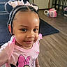 child, girl, braids, pink_bows, minnie_mouse, tshirt, smile, indoor, living_room, wooden_floor, toy, earrings, cute, person, portrait, young_child, fashion, happy, home, floor
