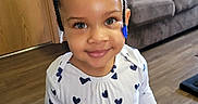 Mi'Chylah is registered to the contest to win money with this photo: toddler, child, smiling, braids, hair_bows, indoor, wooden_floor, white_shirt, heart_pattern, blue_hearts, holding_object, black_clip, living_room, sofa, cushion, door, happy, cute, portrait, person