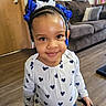 toddler, child, smiling, braids, hair_bows, indoor, wooden_floor, white_shirt, heart_pattern, blue_hearts, holding_object, black_clip, living_room, sofa, cushion, door, happy, cute, portrait, person