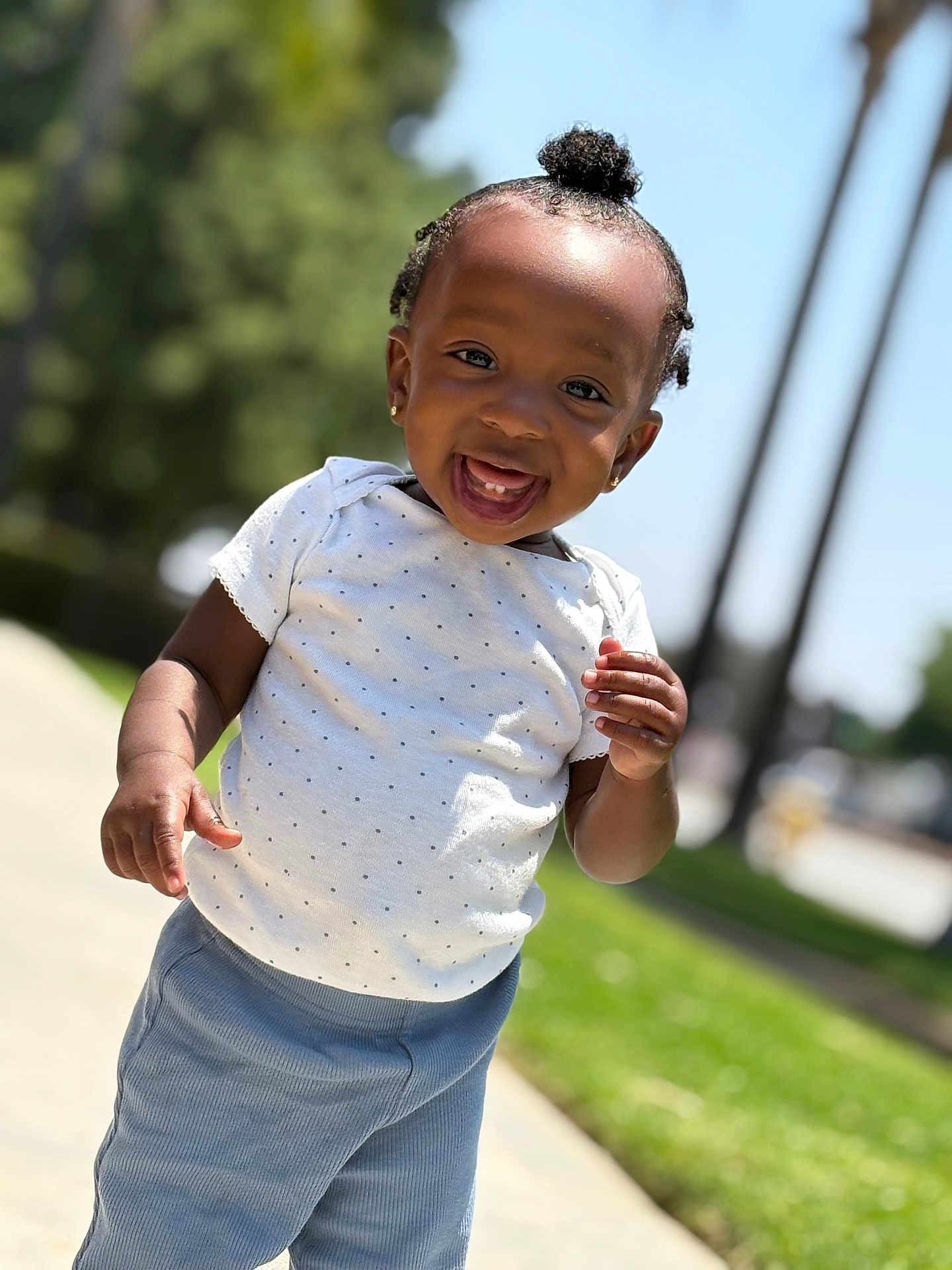 Cali is registered to the contest to win money with this photo: toddler, child, smiling, happy, outdoor, daylight, grass, trees, pants, shirt, polka_dot, hairstyle, top_knot, person, portrait, standing, sidewalk, sunlight, cute, young_child
