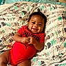 Kyzon is registered to the contest to win money with this photo: baby, child, infant, smiling, happy, lying_down, bed, blanket, bedroom, red_outfit, onesie, portrait, face, hands, arms, legs, socks, patterned_bedding, indoor, cozy