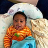 baby, infant, child, smiling, happy, portrait, lying_down, blanket, blue_blanket, orange_clothing, striped_clothing, cushion, bedding, indoors, home, nursery, patterned_fabric, dark_hair, open_mouth, cozy