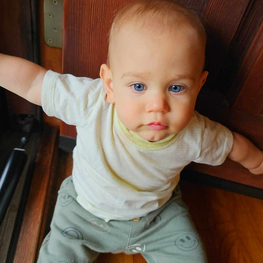 Hayes joined the competition — help win amazing prizes! baby, child, infant, portrait, sitting, blue_eyes, blond_hair, white_shirt, gray_pants, smiley_faces, wooden_floor, wooden_door, doorway, indoor, home, lighting, close_up, looking_at_camera, outstretched_arms, cute